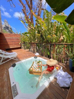 Un jacuzzi con una bandeja de comida y bebidas, rodeado de abundante follaje y una terraza de madera.