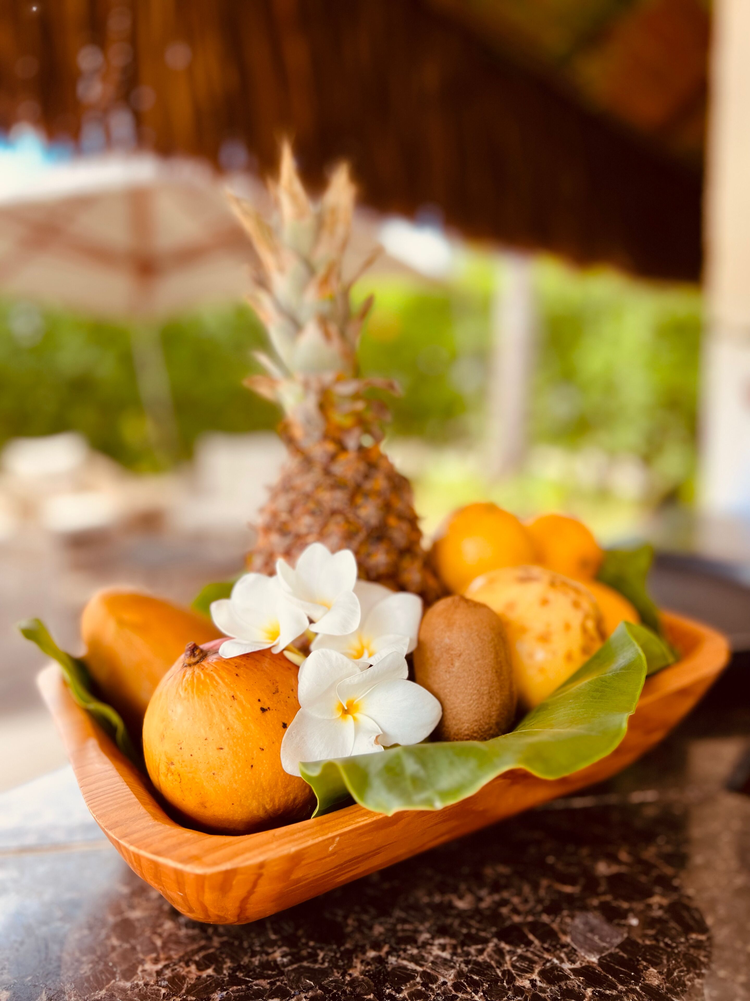A wooden bowl containing a pineapple, oranges, and kiwis on a marble surface.