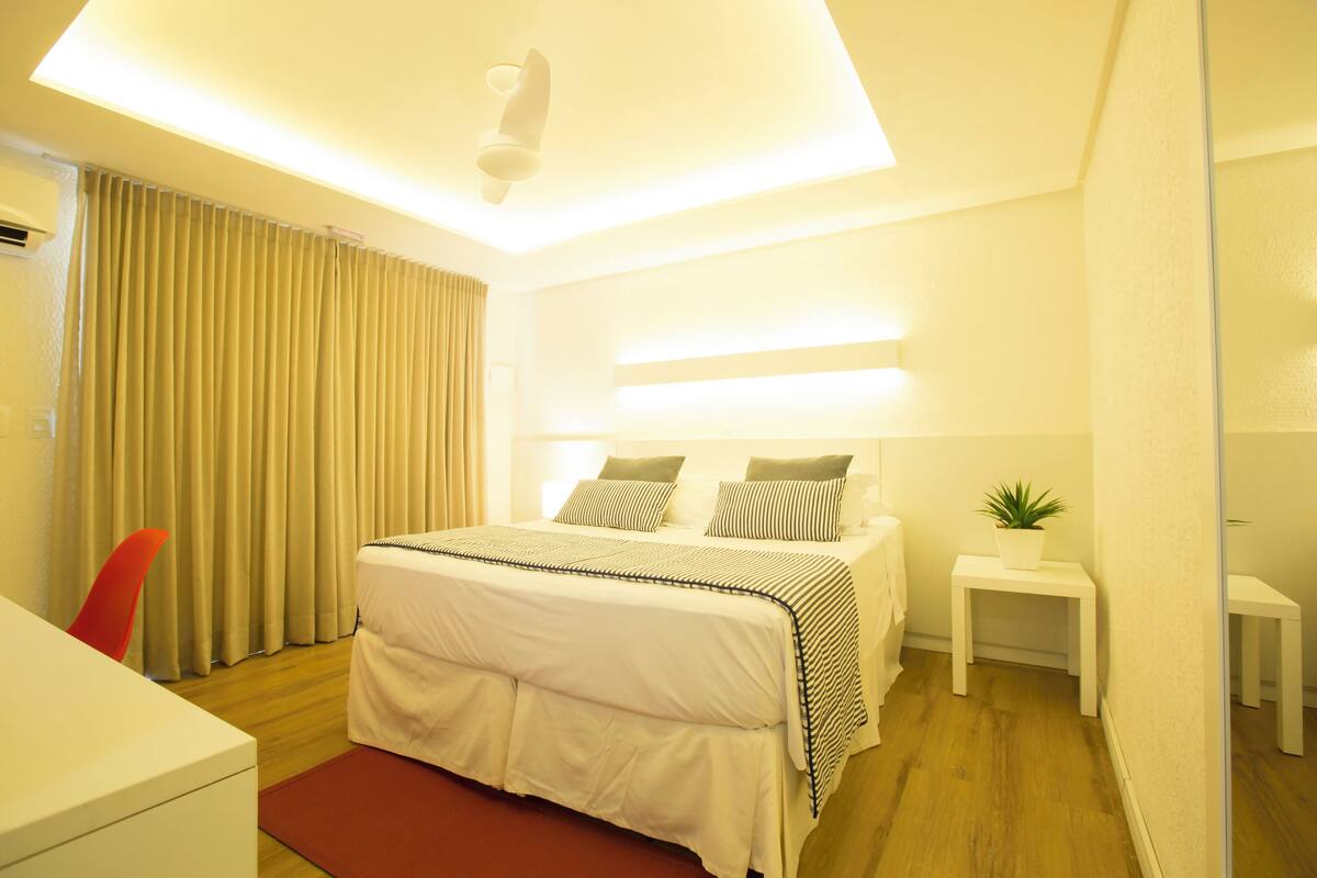 Quarto de hotel moderno com cama, mesas de cabeceira, uma cadeira vermelha e uma pequena planta.