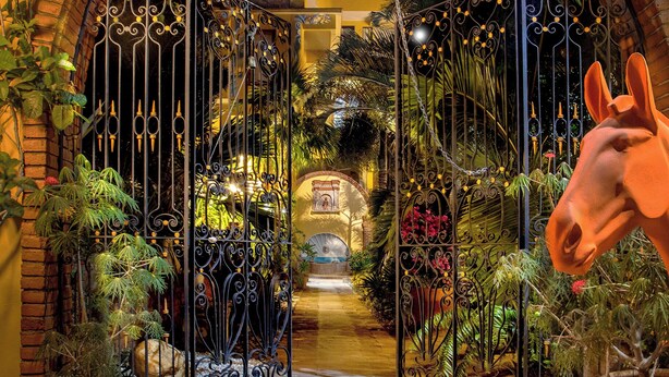 Un portail en fer forgé ouvragé, surmonté d’une arche en briques, mène à un jardin luxuriant.