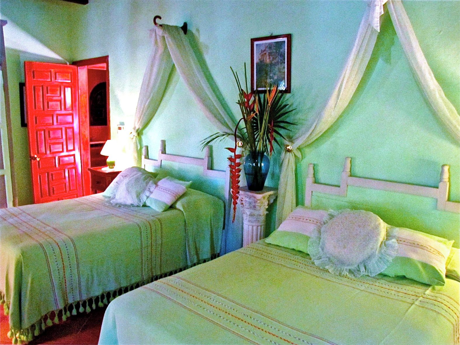 Una habitación con dos camas, una puerta roja, una mesita de noche con una lámpara y un cuadro en la pared.