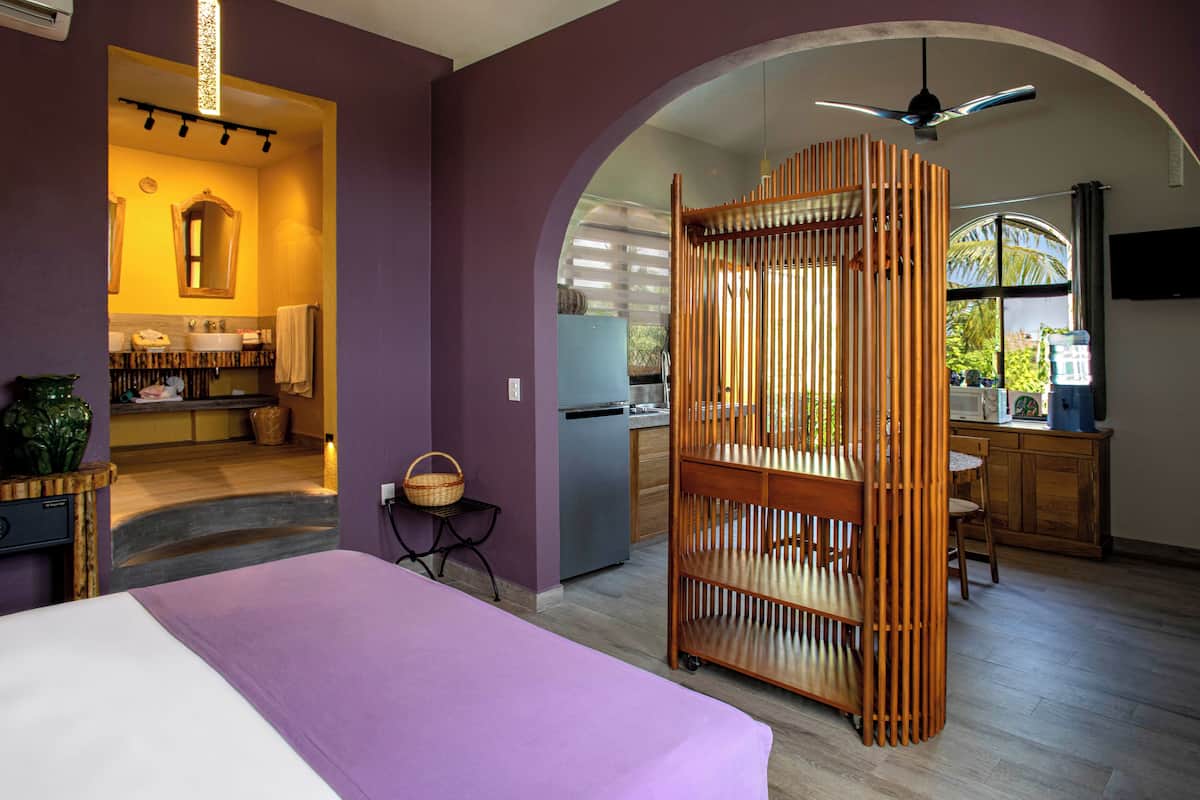 Une pièce comprenant un lit, une cloison en bois, un coin cuisine et une salle de bain avec un miroir et un lavabo.