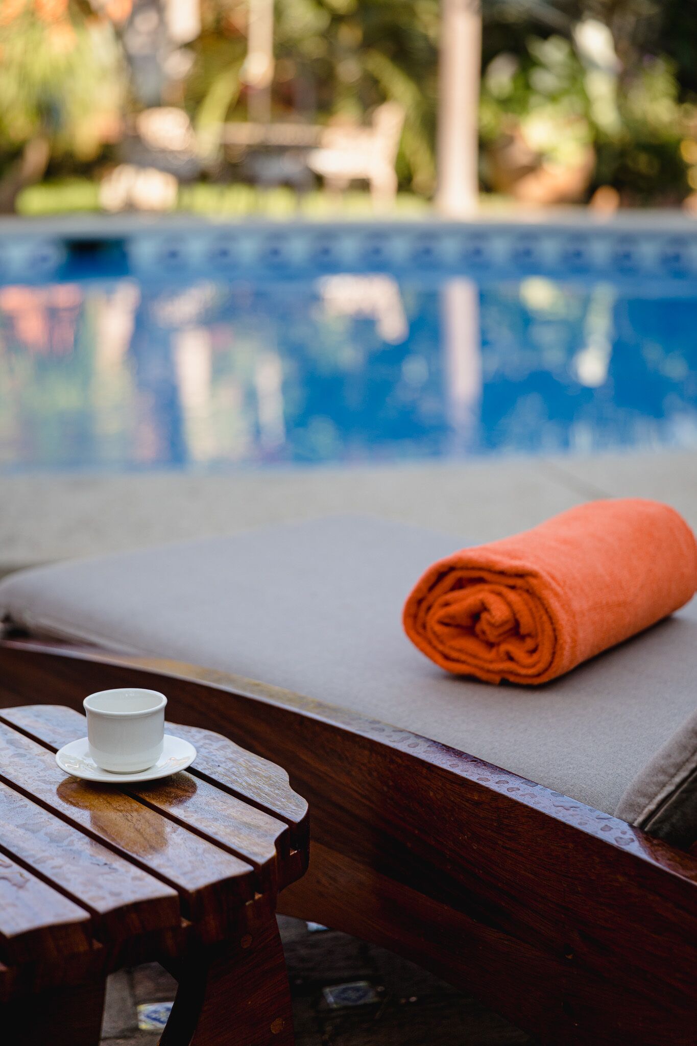 Un banco de madera con una taza y platillo blancos, una toalla naranja enrollada y una piscina al fondo.