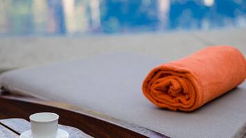 Un banco de madera con una taza y platillo blancos, una toalla naranja enrollada y una piscina al fondo.