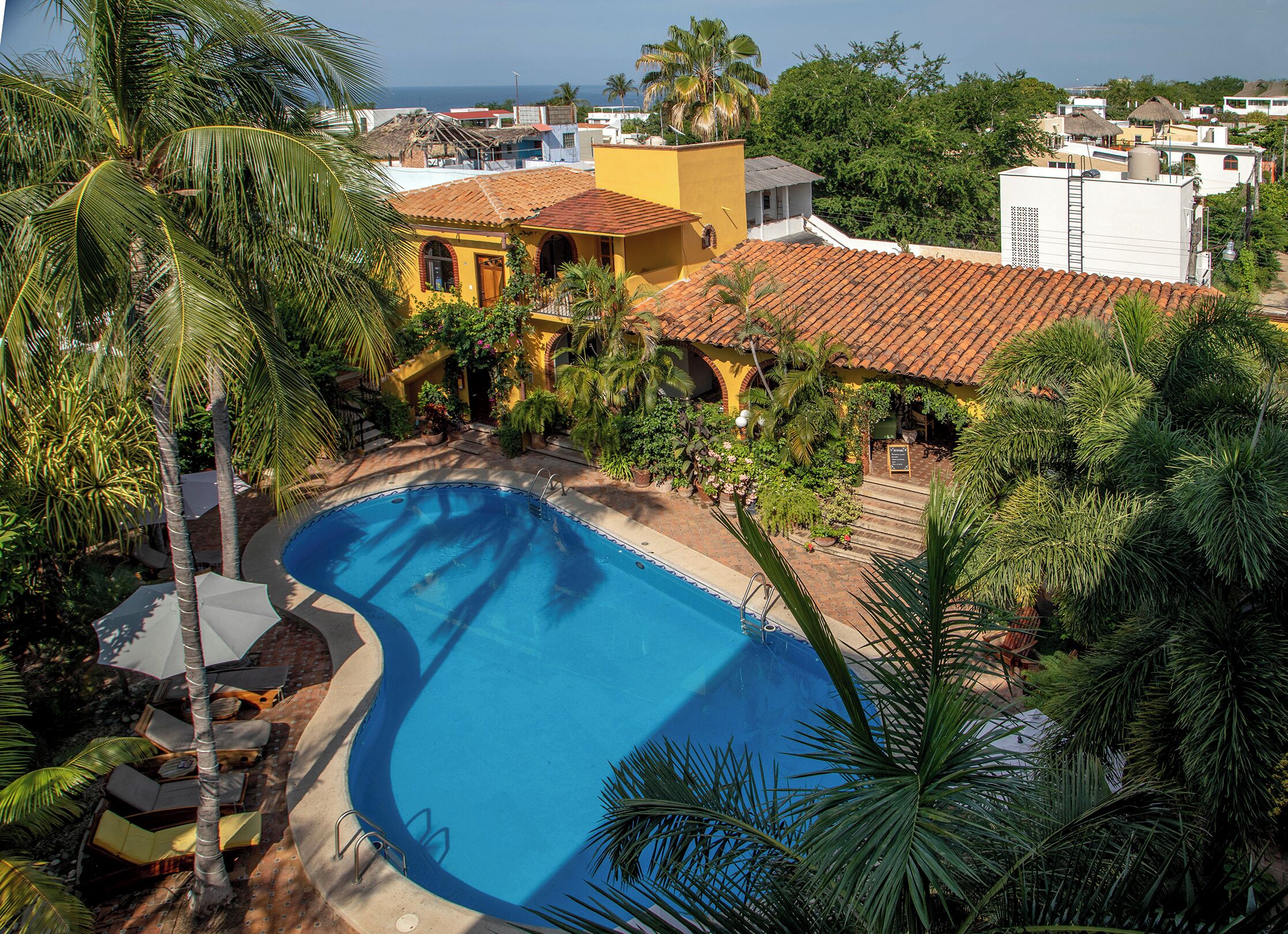 A pool surrounded by lush greenery and a yellow building with a red-tiled roof.
