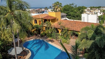 A pool surrounded by lush greenery and a yellow building with a red-tiled roof.