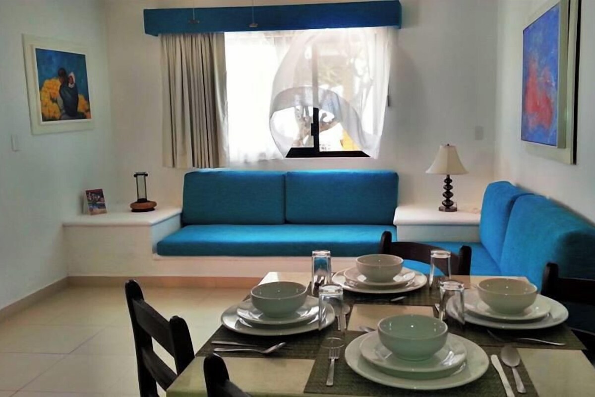 A dining area with a table set for four, a blue sofa, a window with sheer curtains, and artwork on the walls.