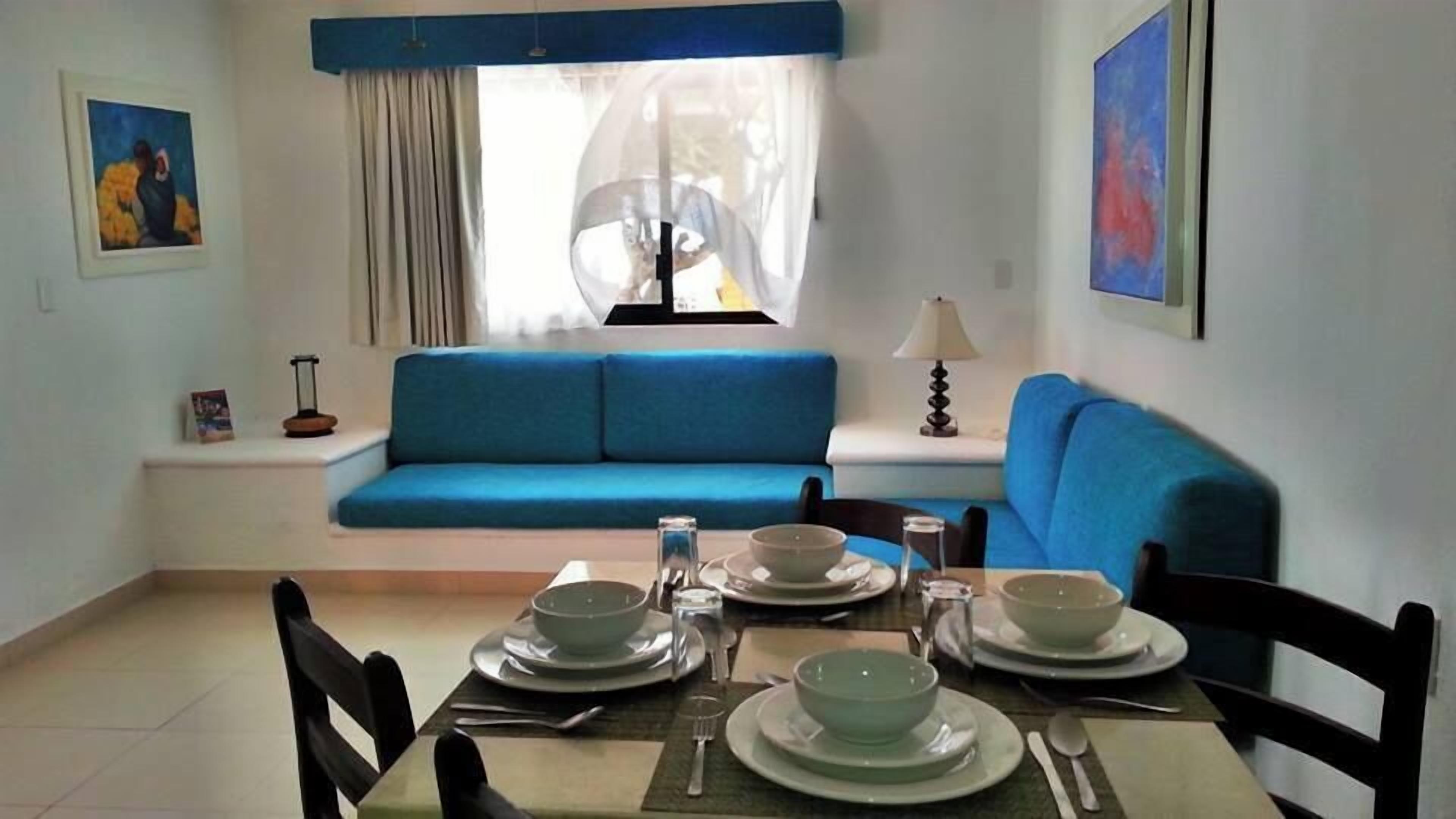 A dining area with a table set for four, a blue sofa, a window with sheer curtains, and artwork on the walls.