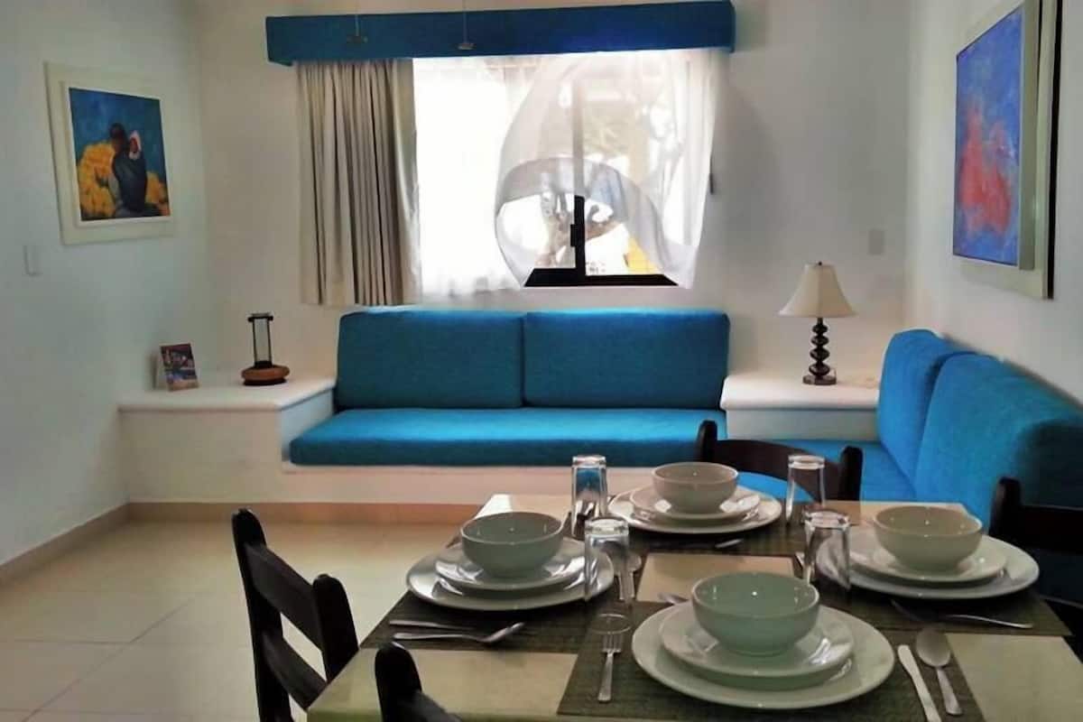 A dining area with a table set for four, a blue sofa, a window with sheer curtains, and artwork on the walls.