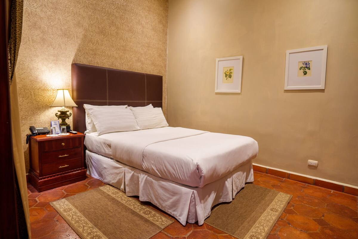 Un lit bien fait, avec du linge de lit blanc, une table de chevet en bois sur laquelle se trouve une lampe, et deux tableaux encadrés accrochés au mur.