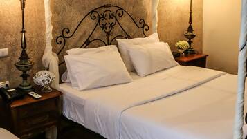 Habitación de hotel con dos camas, cada una con ropa de cama blanca y cabeceros metálicos. Se aprecia una mesita de noche con lámpara, teléfono y un florero con flores. La habitación tiene un papel tapiz estampado y una puerta de madera.