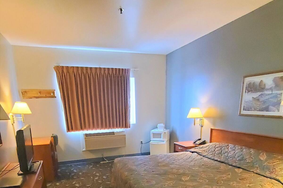 Une chambre d’hôtel comprenant un lit, deux tables de chevet, un bureau, une chaise, une fenêtre avec des rideaux, un tableau au mur et une lampe sur chaque table de chevet.