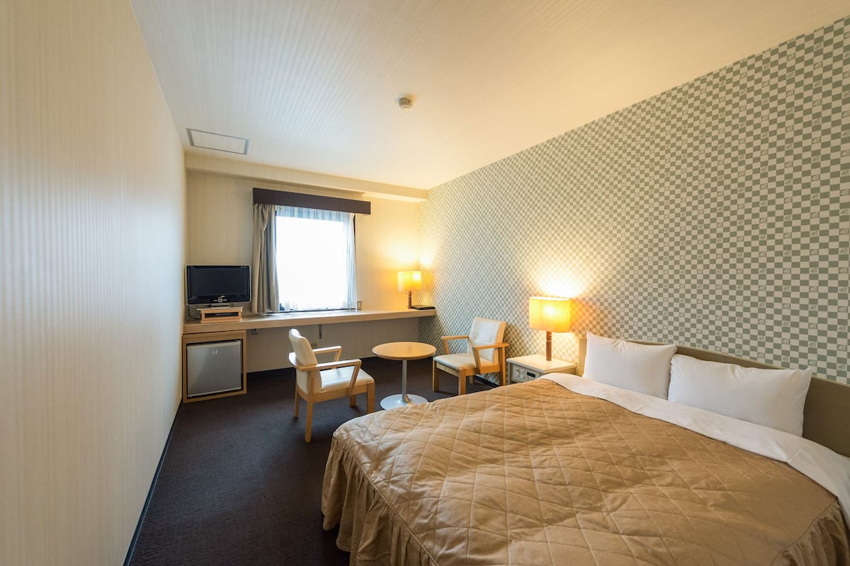 A hotel room with a bed, a desk with a television, two chairs, a small table, and two lamps.