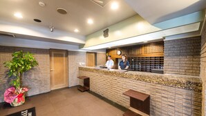 A reception area with a brick wall, a reception desk, a clock, and a potted plant. - Hotel LiVEMAX Kofu (Kai)