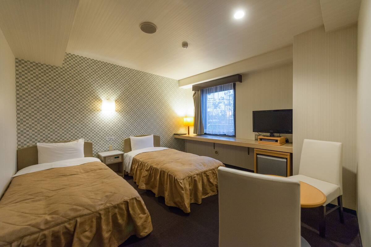 A hotel room with two beds, a desk with a television, a chair, and a window with curtains.
