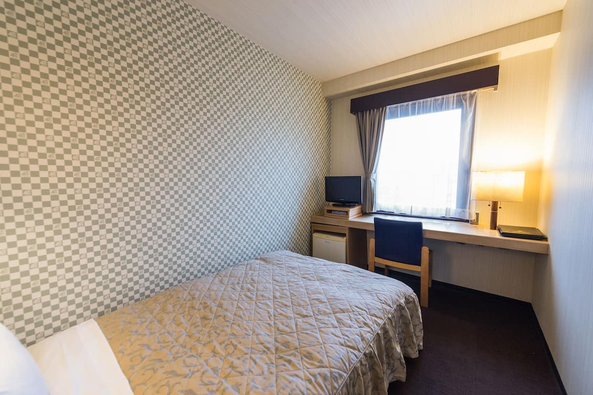 A hotel room with a checkered wall, a bed with a patterned bedspread, a desk with a computer, and a window with curtains.