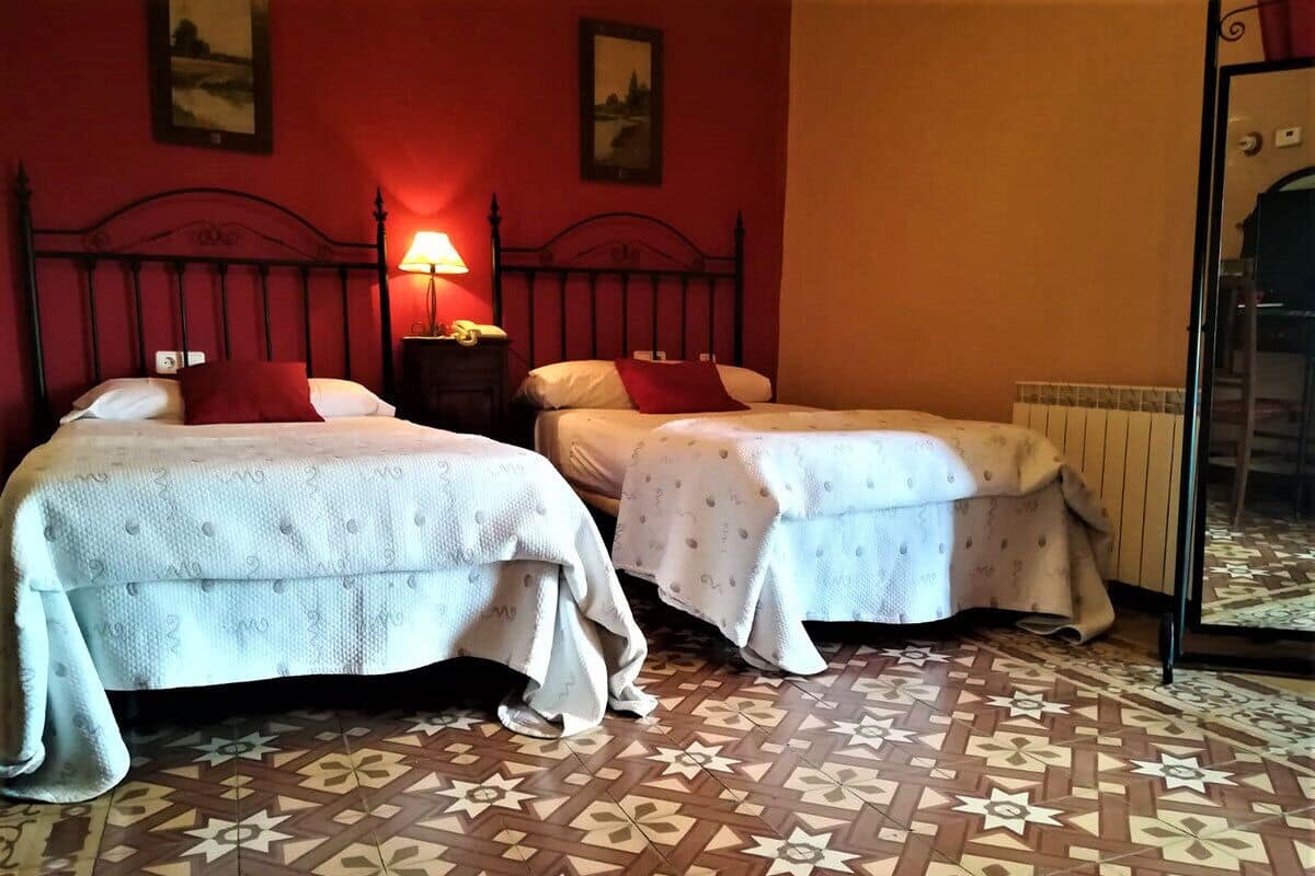 Duas camas de solteiro com cabeceiras, uma mesa de cabeceira com abajur e um chão estampado.