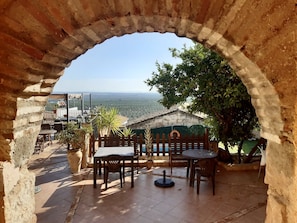 An outdoor patio with a view of a distant landscape, featuring a stone archway, wooden tables, and chairs. - Hotel Palacio Guzmanes (Banos De La Encina)