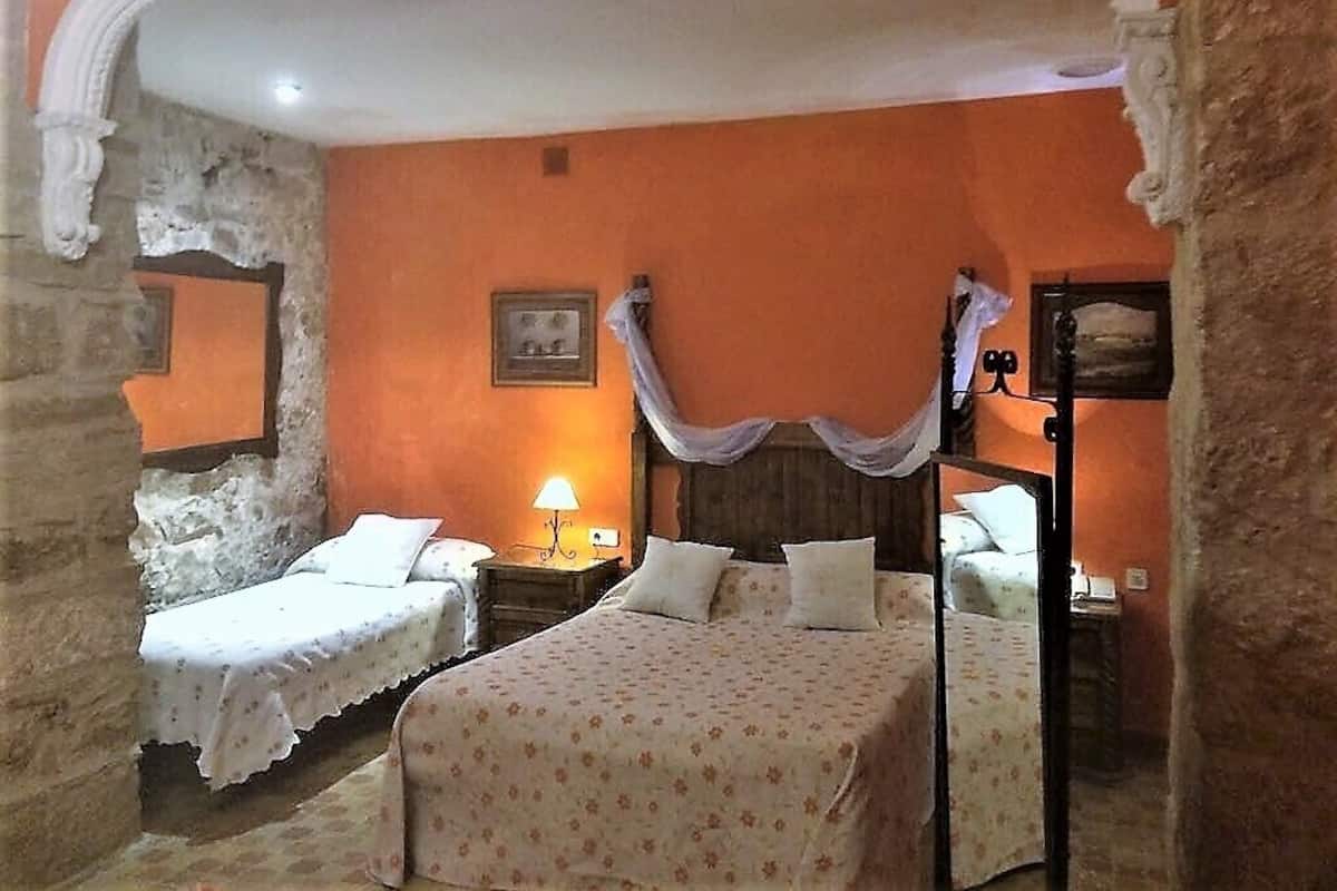 Quarto com duas camas, uma com dossel e outra sem, mesa de cabeceira com abajur e parede de pedra.