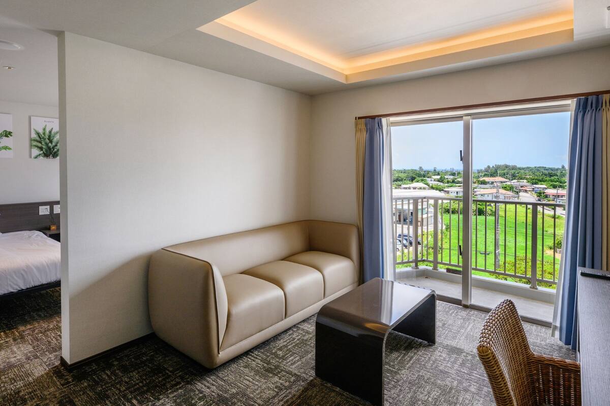 A hotel room with a bed, a sofa, a small table, a chair, and a large window offering a view of the city.