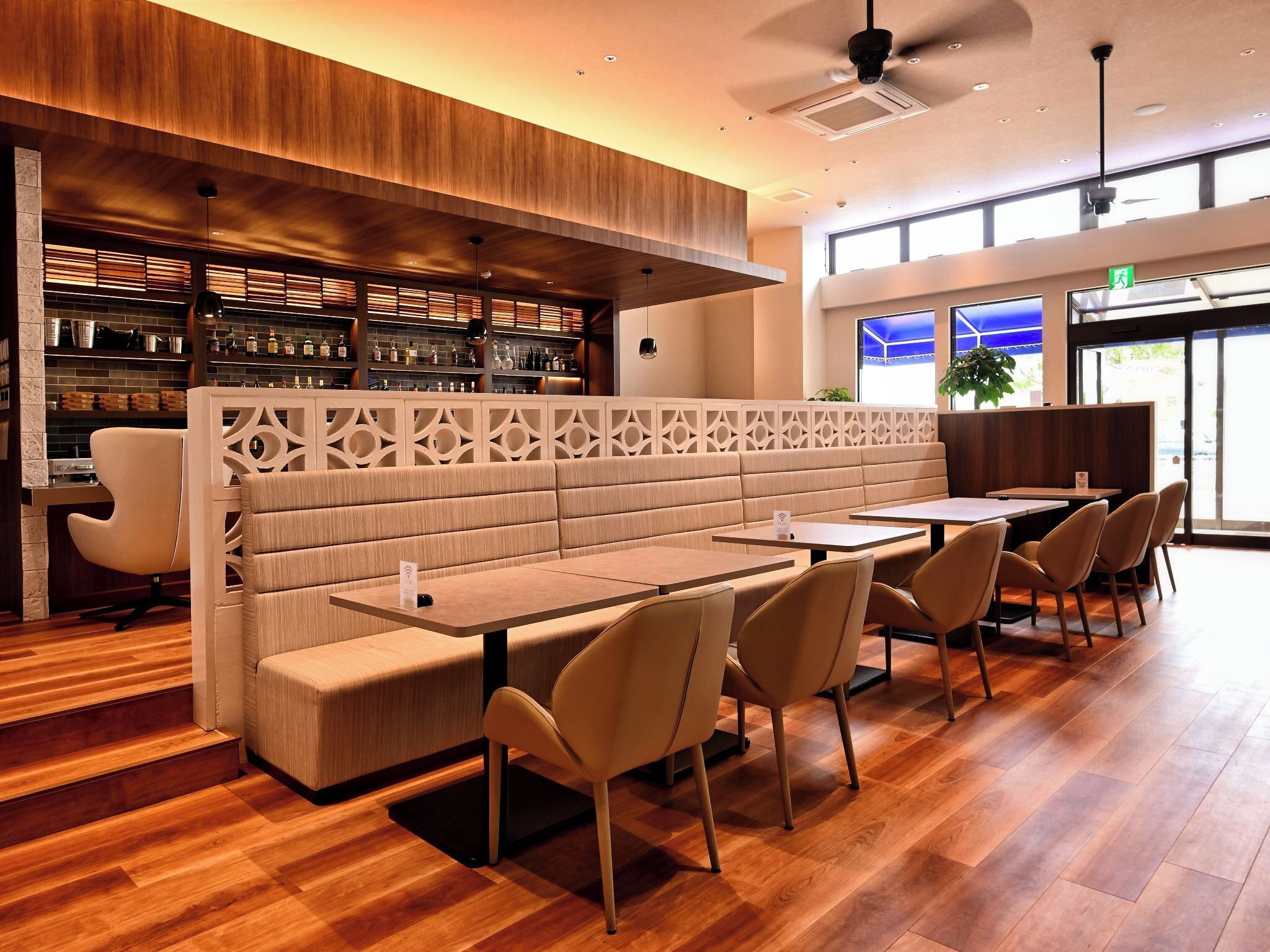 A modern restaurant interior with a wooden ceiling, a bar area with shelves of bottles, and a seating area with tables and chairs.