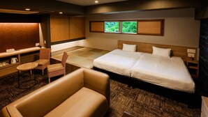 A hotel room with a bed, a seating area with a chair, a small table, a desk, and a window with a view of greenery.
