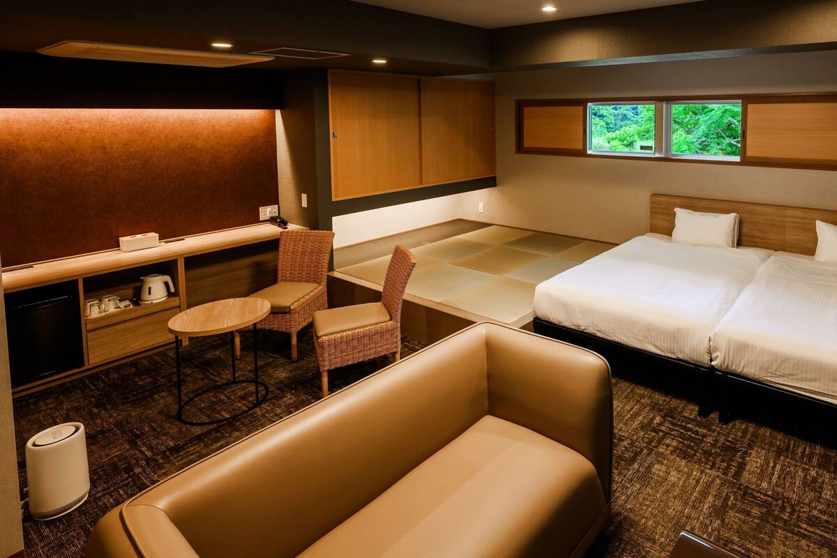 A hotel room with a bed, a seating area with a chair, a small table, a desk, and a window with a view of greenery.