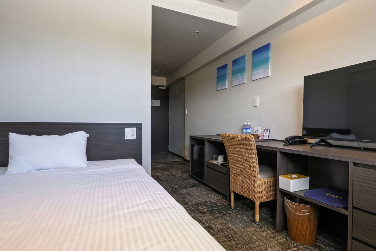 A hotel room with a bed, a desk, a chair, a TV, and a wall-mounted shelf.