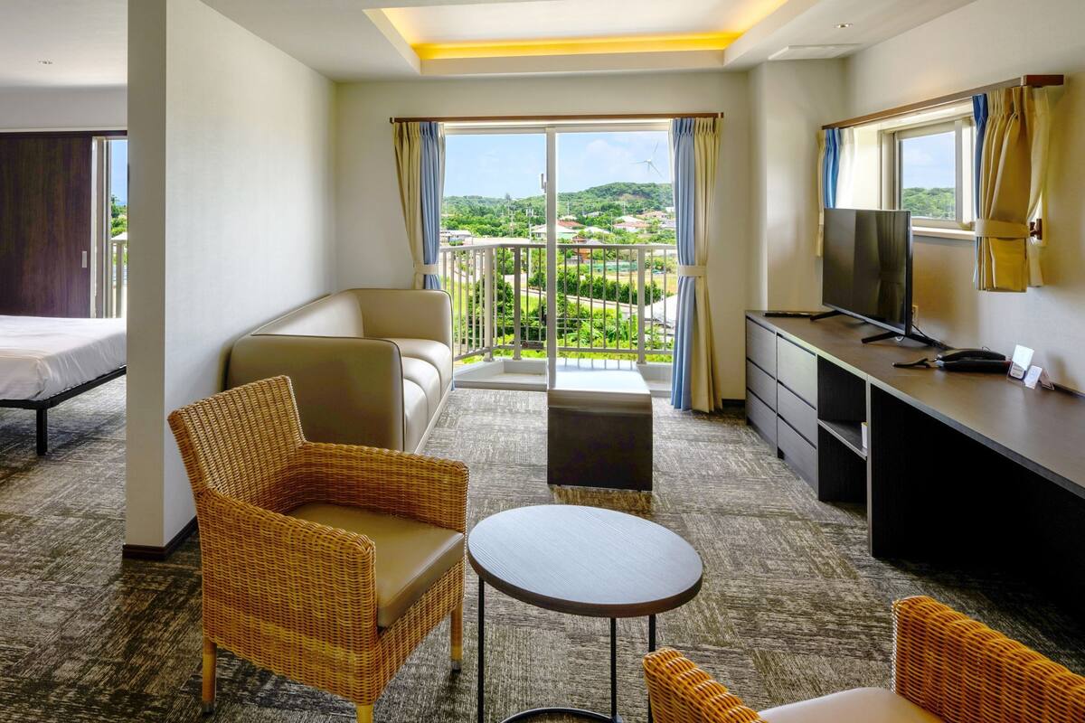 A hotel room with two beds, a sofa, a small table, a TV, and a balcony with a view of the countryside.