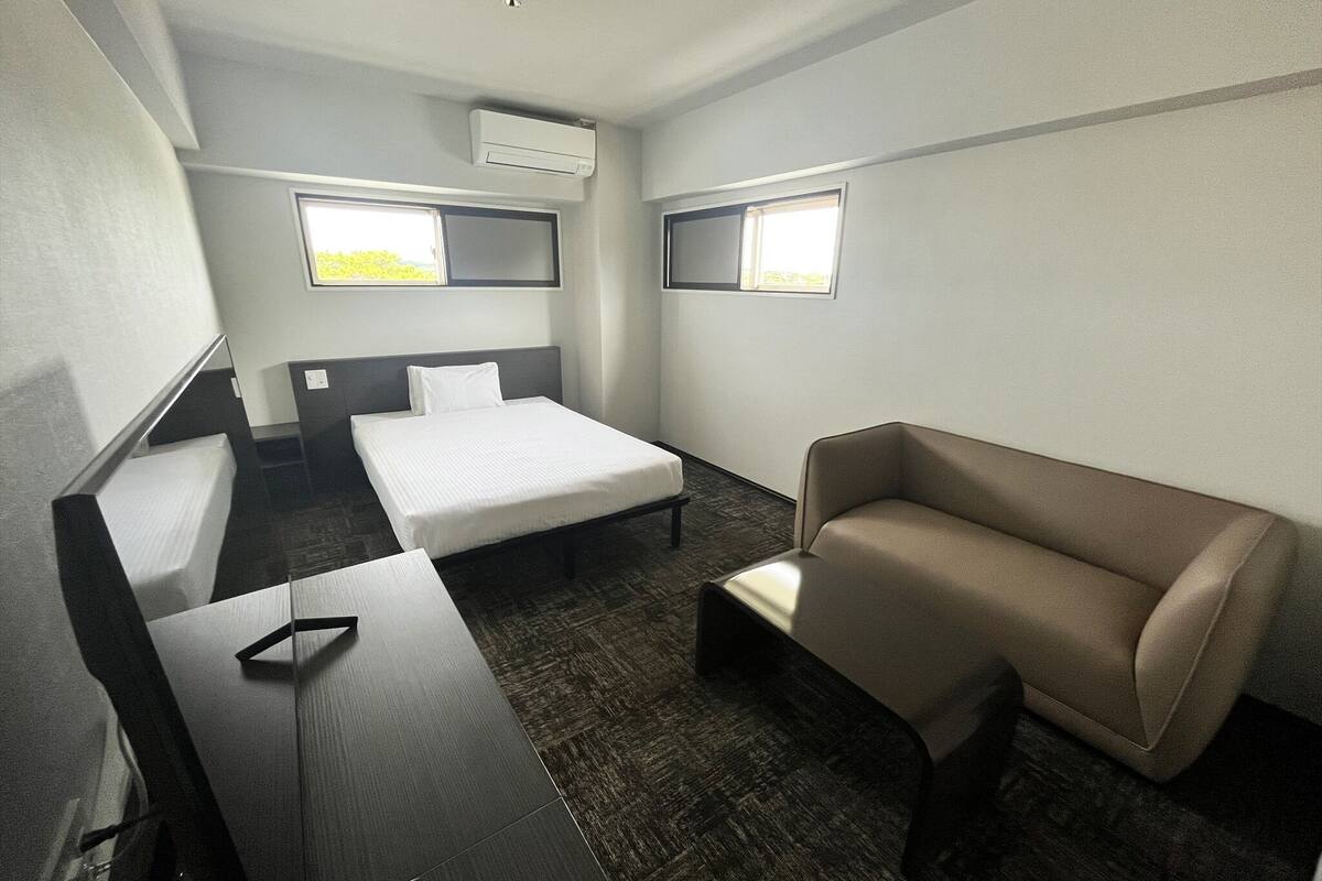 A hotel room with a bed, a sofa, a desk, and two windows.