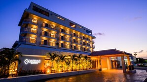 A multi-story hotel with a modern design, featuring balconies on each floor and a well-lit entrance area with palm trees. - Lit Hotel NAKIJIN (Nakijin)