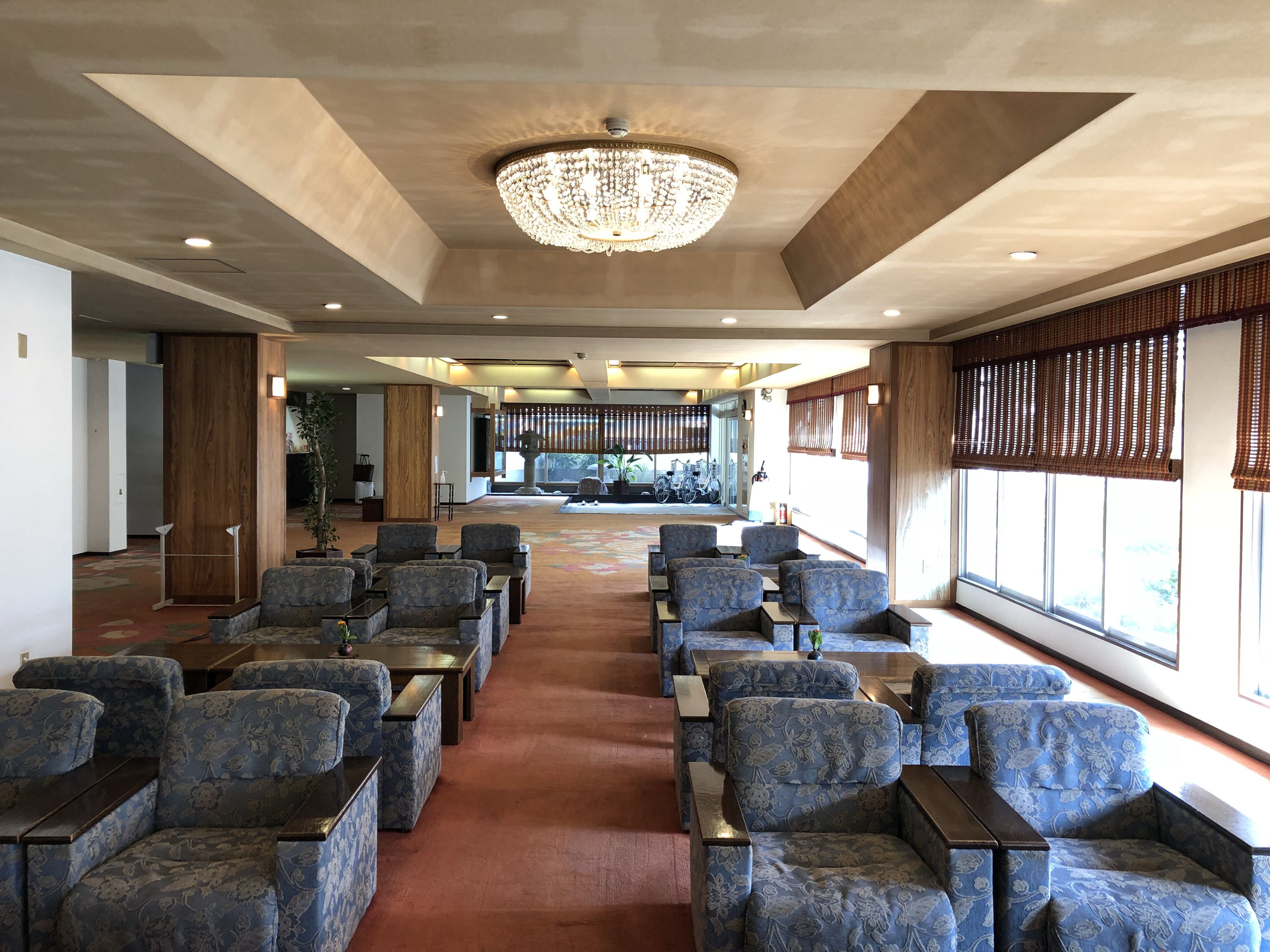 A spacious lobby with rows of blue upholstered chairs and tables, a large chandelier, and large windows with blinds.