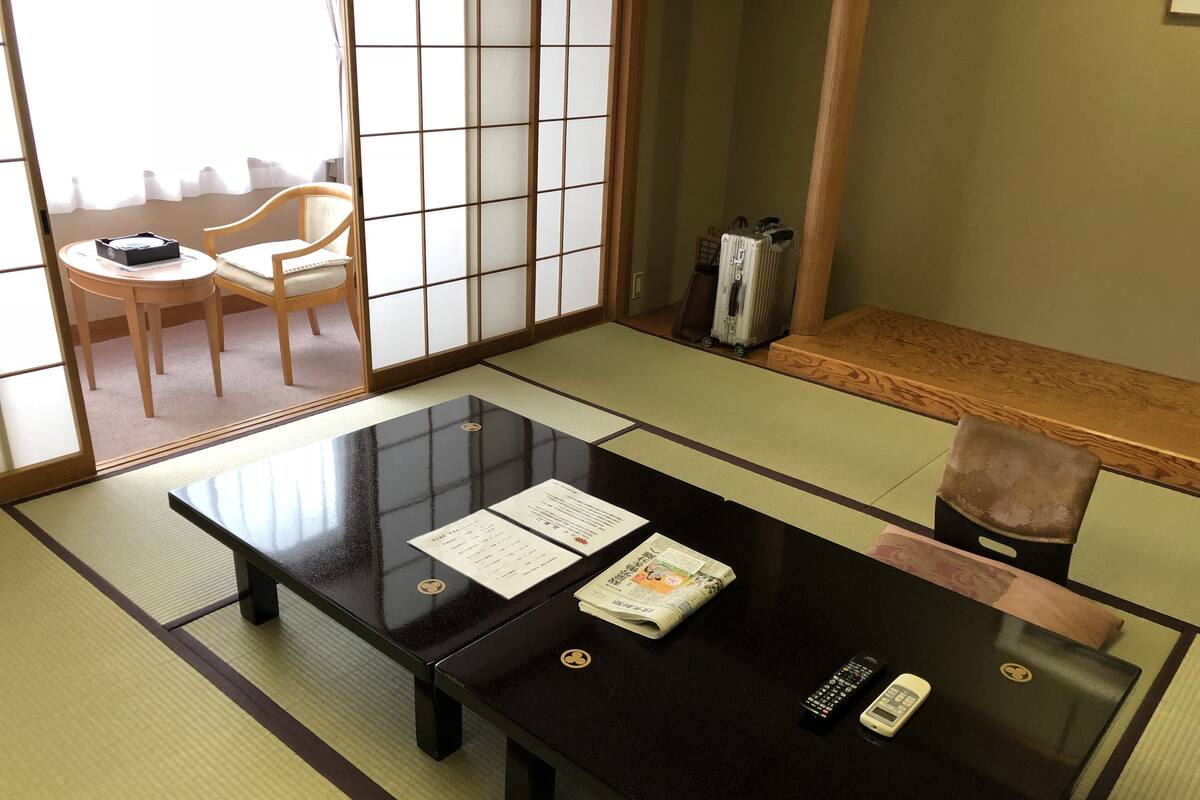 Une pièce de style japonais traditionnel, avec un sol recouvert de tatamis, une table laquée noire sur laquelle se trouvent des documents, une télécommande et une chaise.
