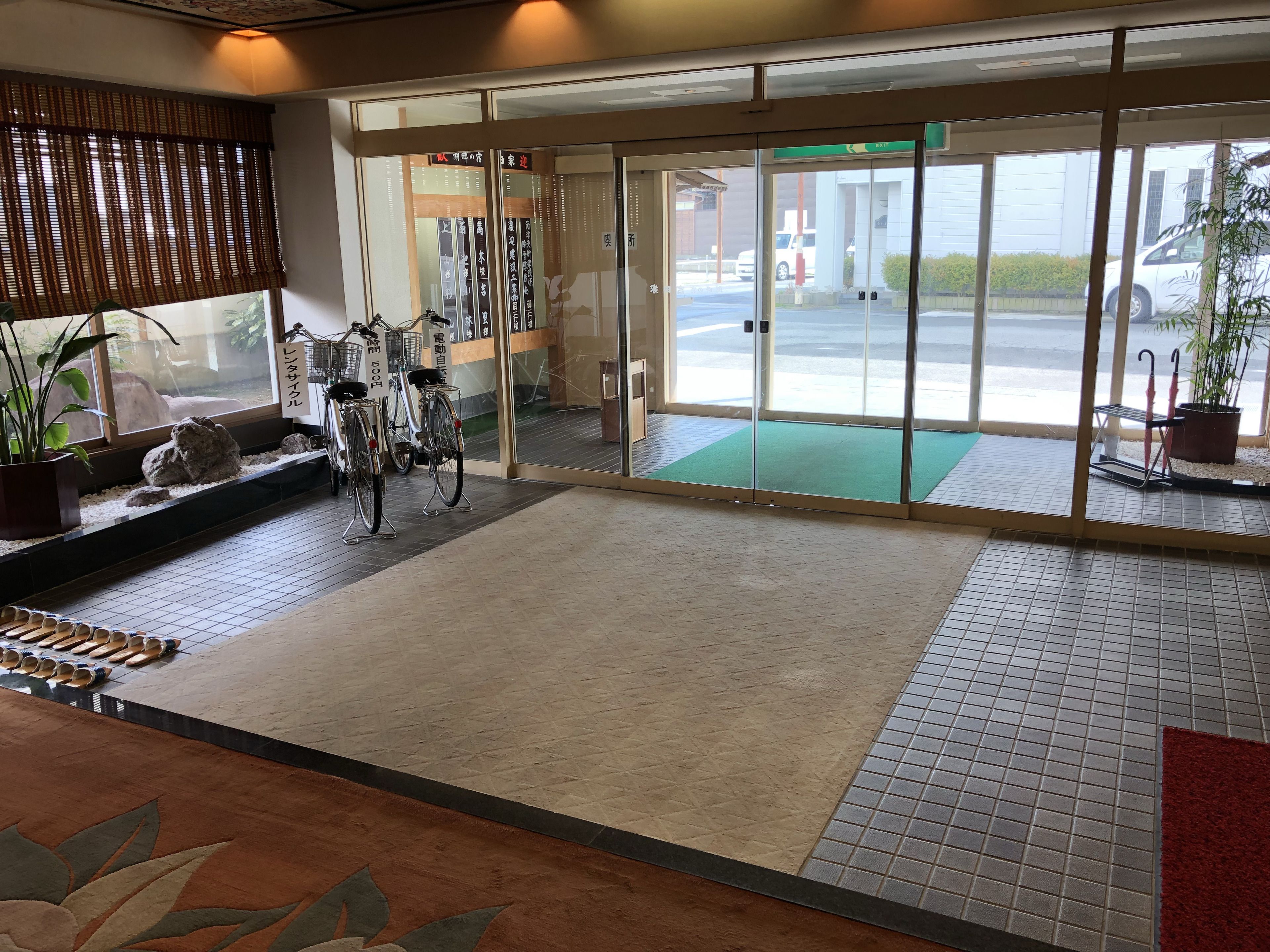 Un hall d’hôtel avec un sol carrelé, un comptoir d’accueil, des vélos et une vue sur le parking à travers des portes vitrées.
