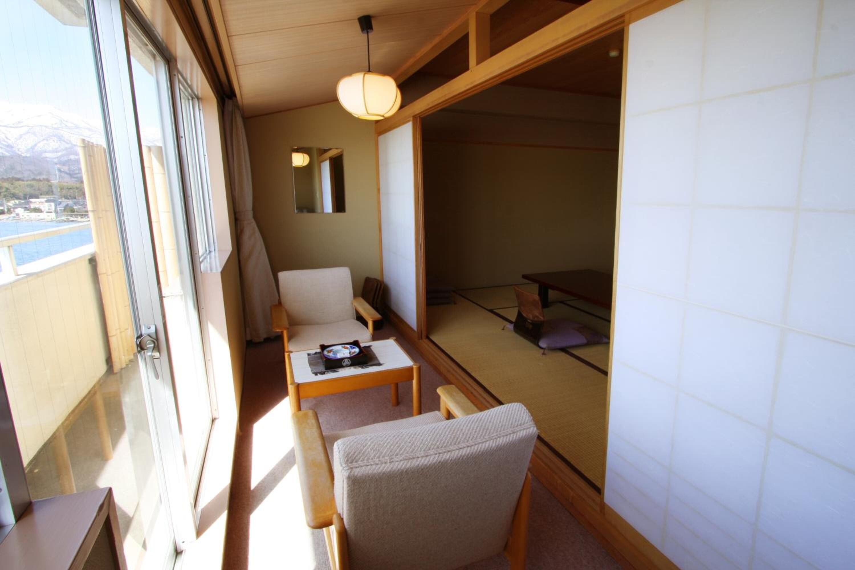 Una habitación tradicional japonesa con suelo de tatami, una mesa baja, una silla y una puerta corredera que da a un balcón con vistas a montañas.