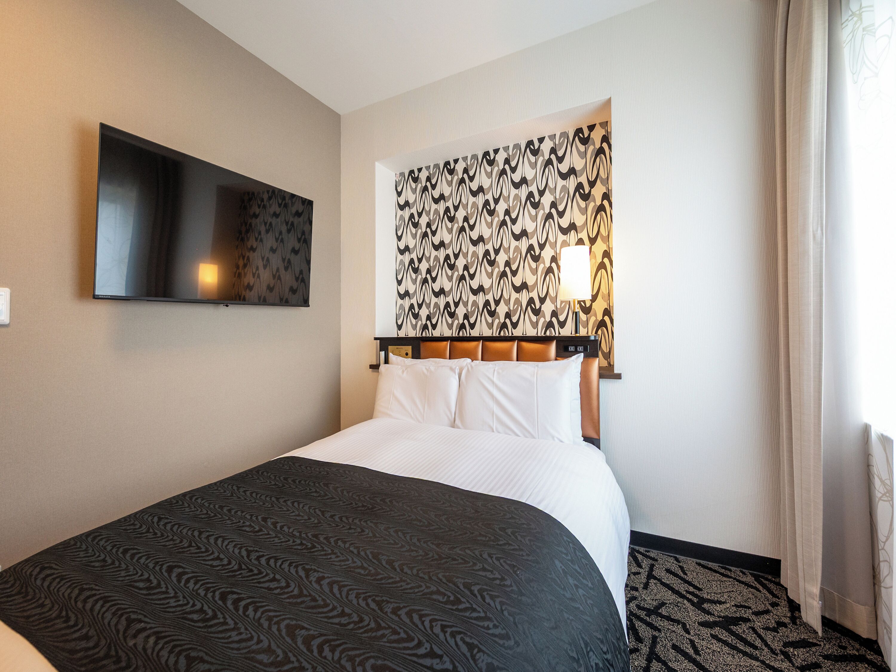 Une chambre d’hôtel avec un lit, un téléviseur à écran plat fixé au mur, une tête de lit à motifs et une lampe.