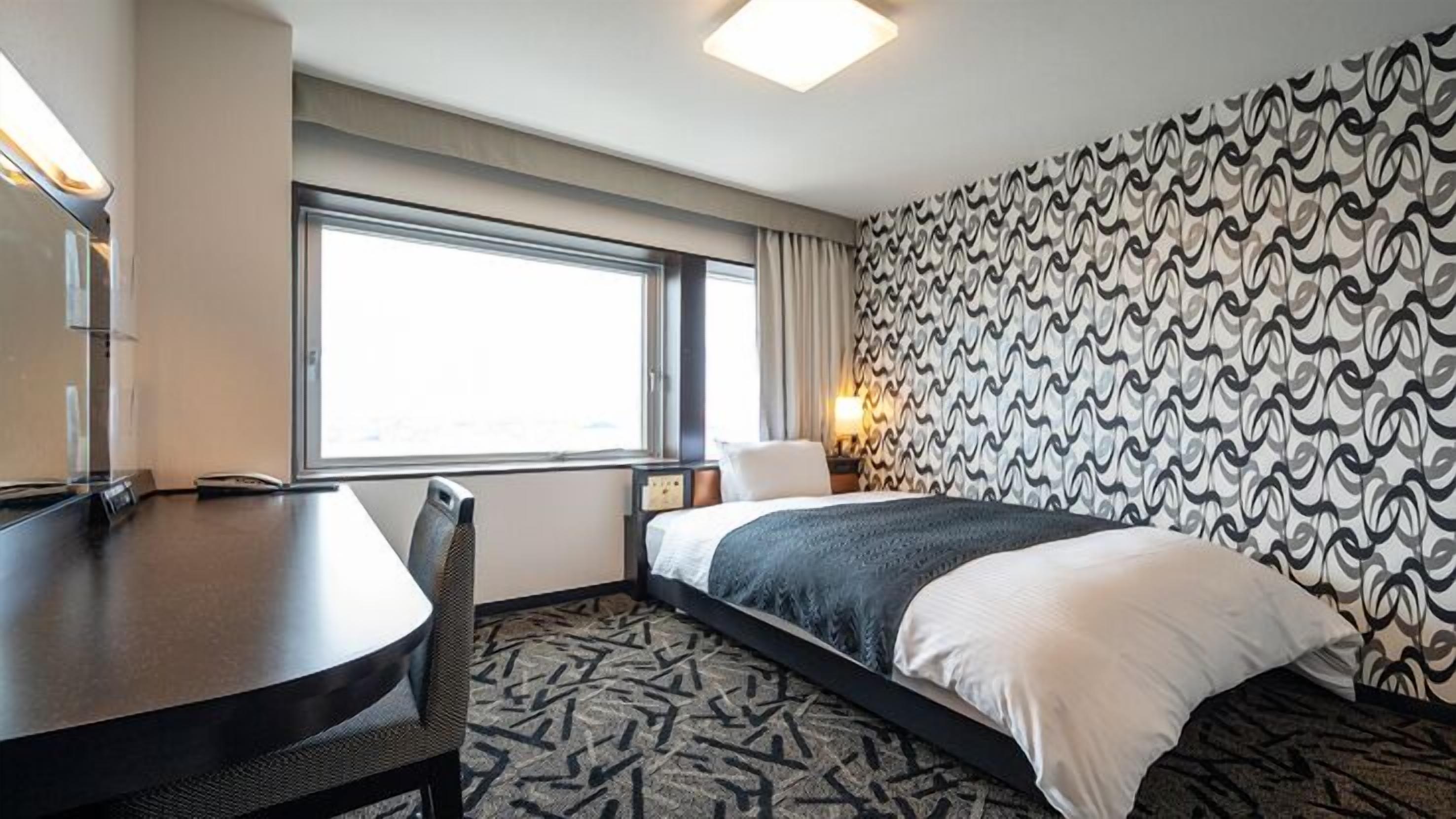 A hotel room with a bed, a desk, a chair, a window, and a patterned wall.