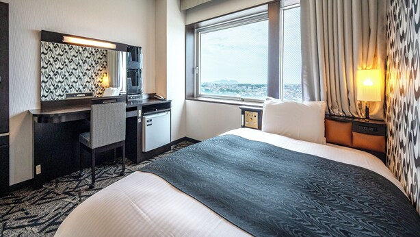 Habitación de hotel con cama, escritorio, silla, espejo, ventana con cortinas y vista a la ciudad.
