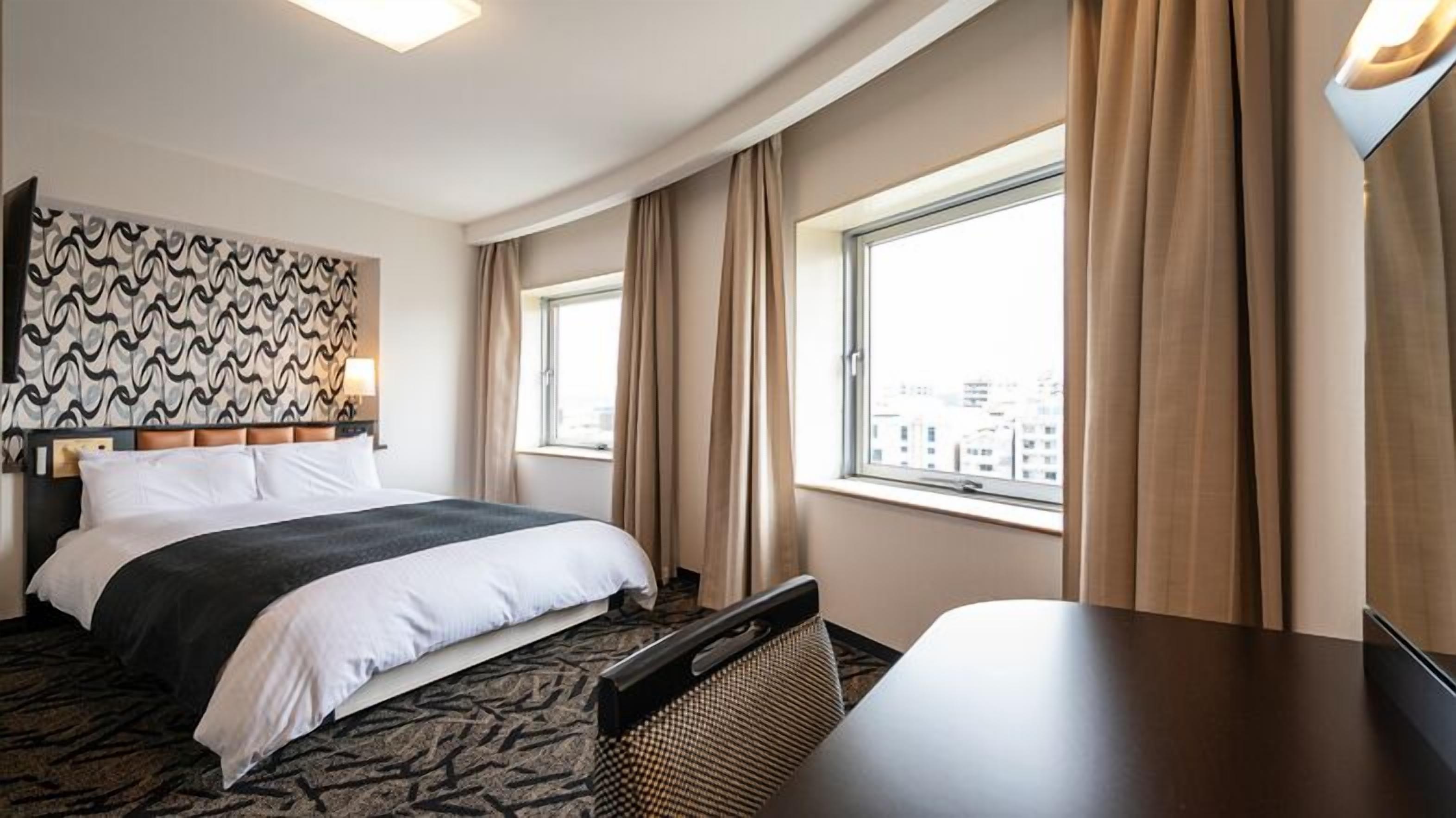 A hotel room with a bed, a desk, a window with curtains, and a city view.