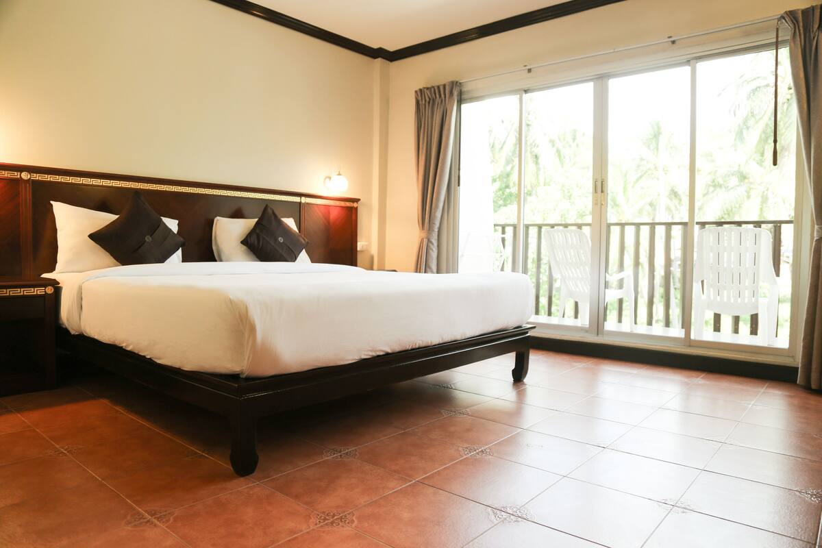Habitación de hotel con una cama grande, una mesita de noche, un ventanal con vistas a palmeras y un balcón con sillas blancas.
