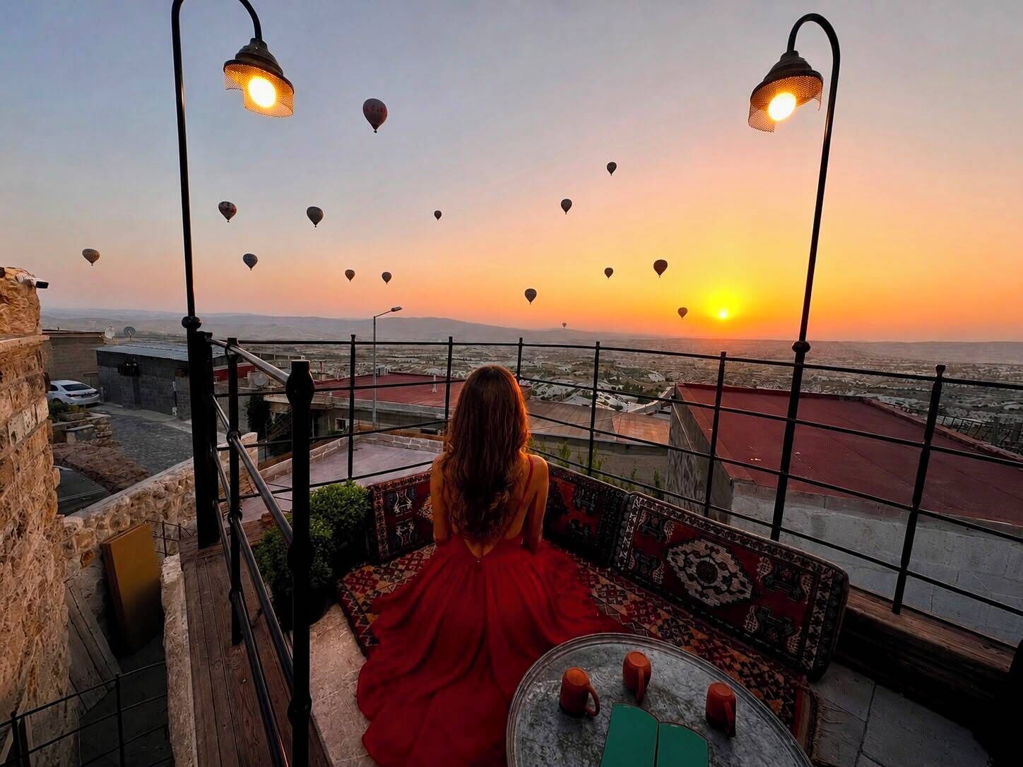 A person sitting on a balcony overlooking a sunset with hot air balloons in the sky.
