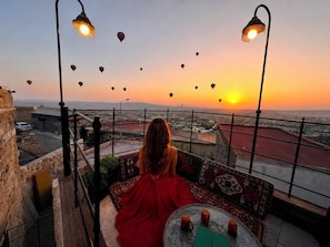 Una persona sentada en un balcón contemplando un atardecer con globos aerostáticos en el cielo.