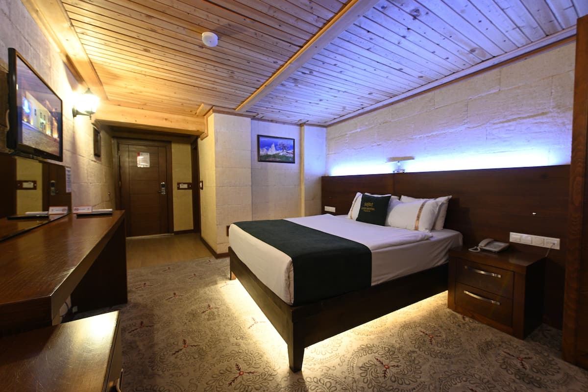 Quarto de hotel com teto de madeira, cama com manta verde, mesa de cabeceira com telefone e televisão montada na parede.