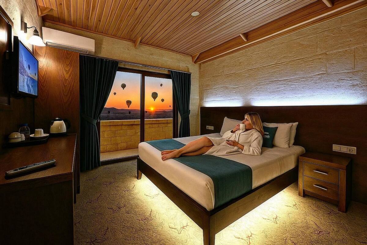 Quarto de hotel com cama, escrivaninha, televisão e vista para balões de ar quente ao pôr do sol.