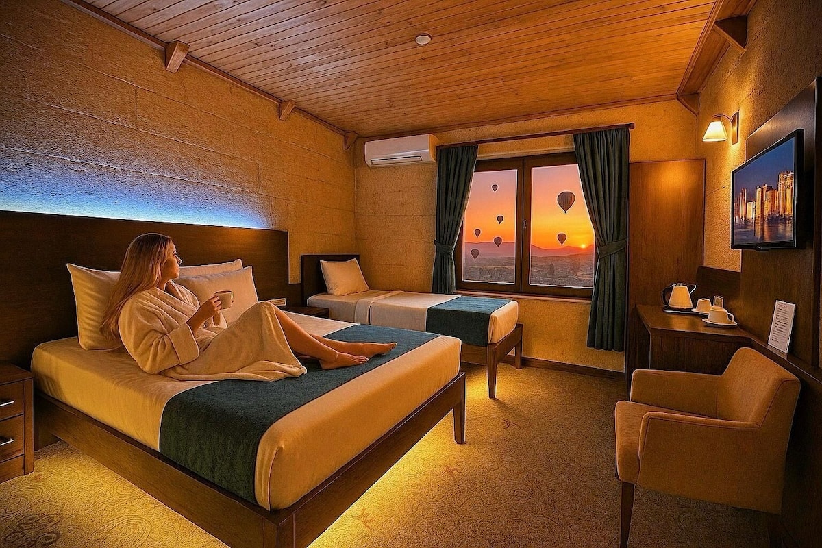Quarto de hotel com cama, sofá, escrivaninha e vista para balões de ar quente ao pôr do sol.