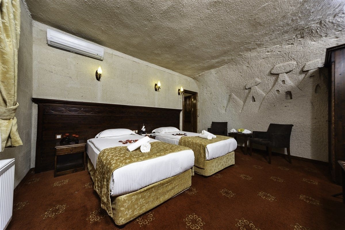 Quarto de hotel com duas camas, cabeceira de madeira, uma mesinha e uma cadeira.
