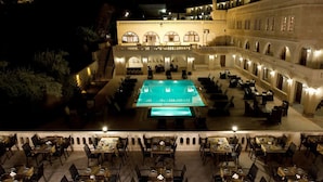 A rooftop pool area with multiple dining tables and chairs, surrounded by a building with arched windows and a staircase leading to an upper level.
