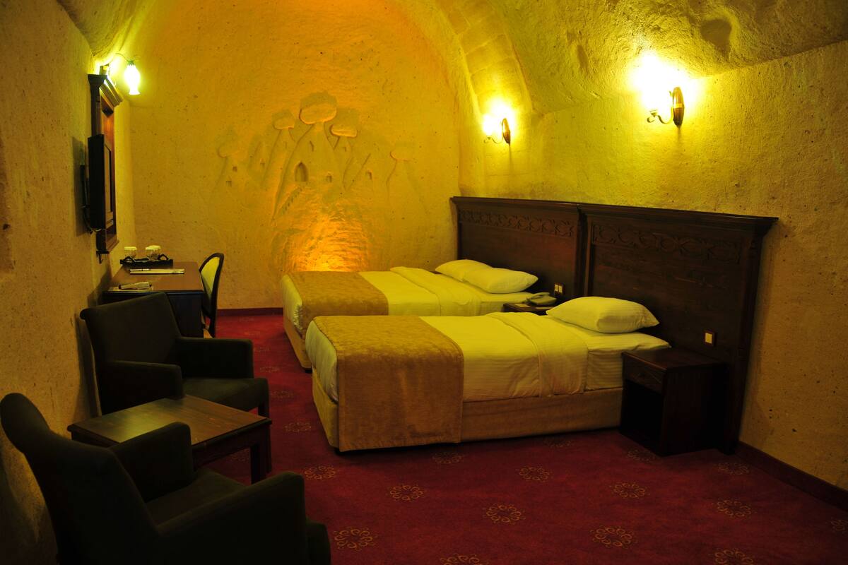 Quarto de hotel com duas camas, uma cabeceira de madeira, uma mesinha, uma cadeira e uma luminária fixada na parede.