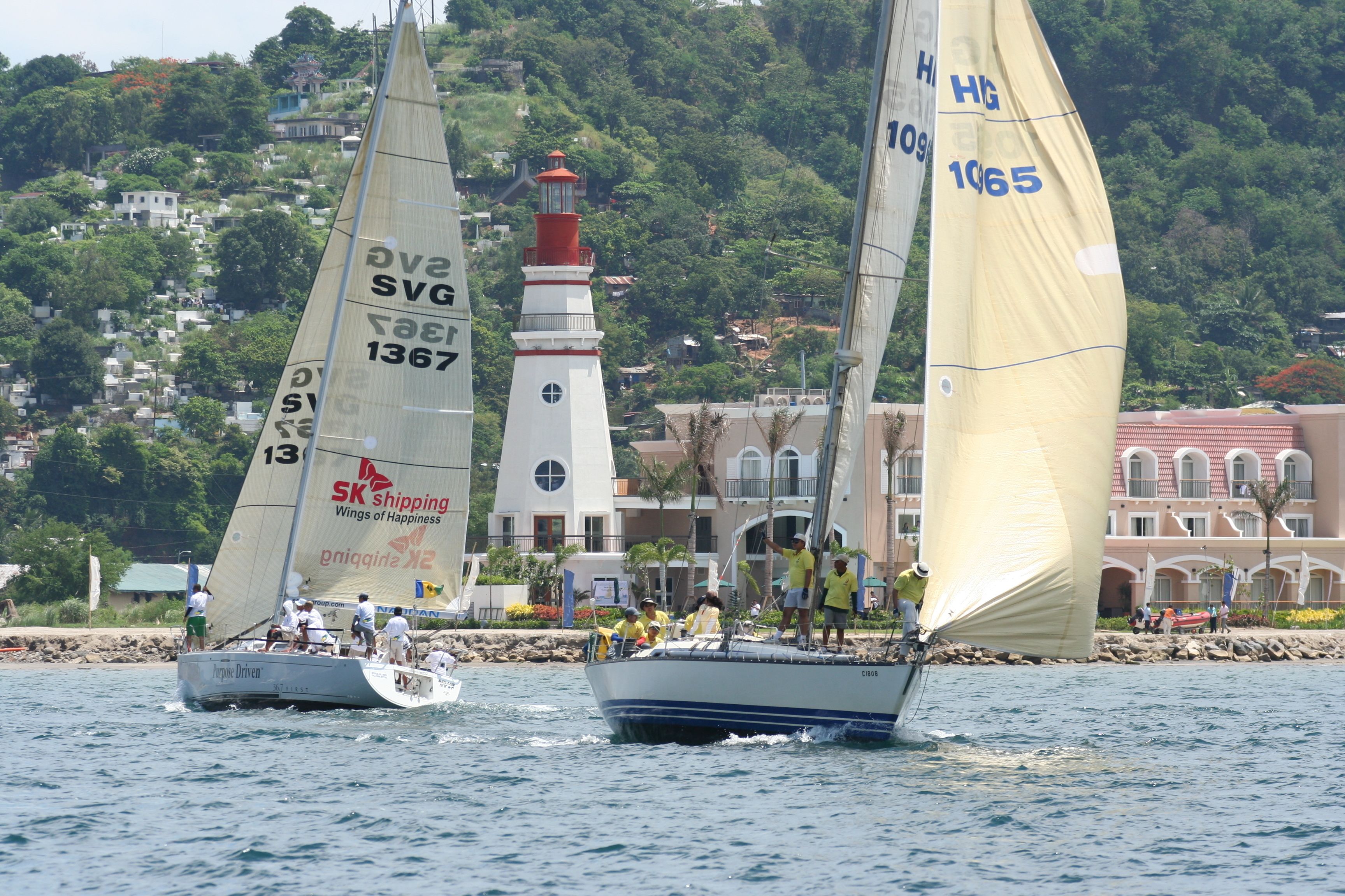 A sailboat with the number 1367 on its sail, sailing near a coastal town with buildings and a lighthouse in the background.