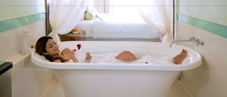 A bathtub with rose petals on the floor, a white sink, and a bed with a nightstand in the background.